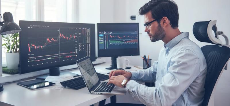 Financial market analyst working at a multi screen trading workstation displaying live market charts, trading data, and technical analysis tools in a modern office environment.