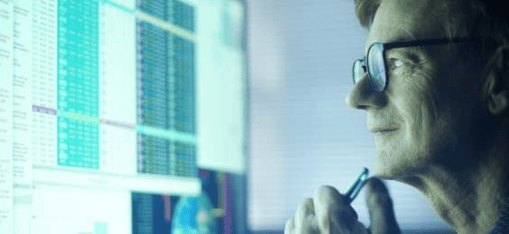 A computer monitor displaying detailed financial data tables and charts in a trading interface, with a person holding a pen while reviewing the information. The setup represents trade scaling and market analysis.