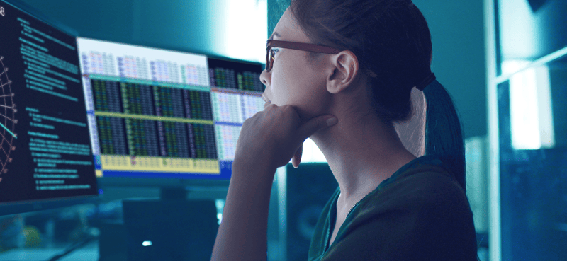 A person seated at a workstation with multiple monitors displaying financial charts, data tables, and technical indicators in a blue-toned office environment. The screens show detailed market analytics and trading information.