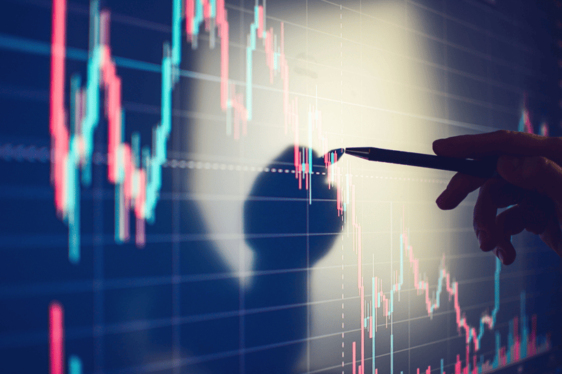 Hand pointing at a large screen displaying colorful candlestick charts and financial data trends