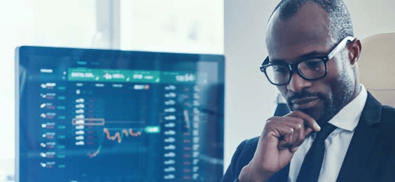 A person in formal attire seated at a desk with a large monitor displaying candlestick charts, trend lines, and market data in a trading interface. The setup represents professional financial analysis and strategy planning.
