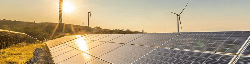 A solar farm with reflective solar panels in the foreground and multiple wind turbines in the background during a warm sunset, showcasing modern renewable energy infrastructure.