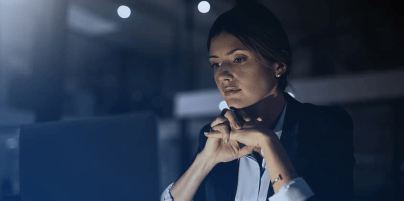 A professional sitting at a desk in a dimly lit office environment with hands clasped near a computer monitor. The scene suggests focus on strategic planning or risk assessment.