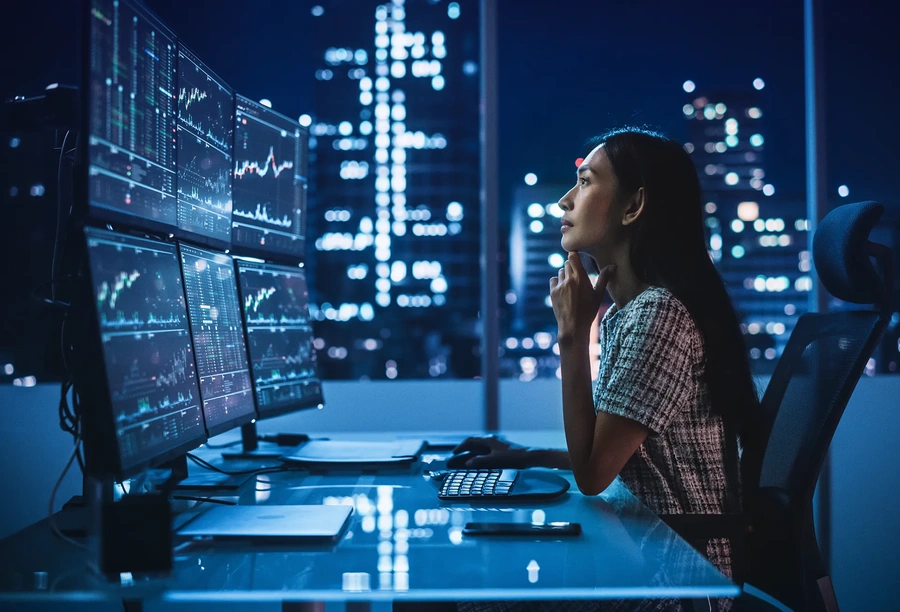 Modern office desk with multiple monitors displaying financial charts and market data in a high rise setting at night.