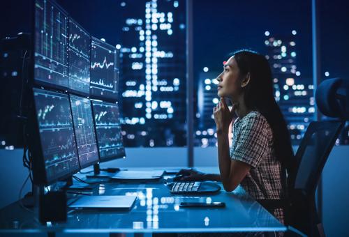 Modern office desk with multiple monitors displaying financial charts and market data in a high rise setting at night.