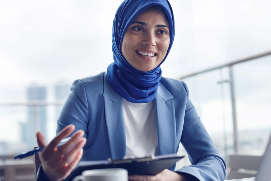 Person wearing a blue headscarf and blue blazer holding a clipboard during a meeting.