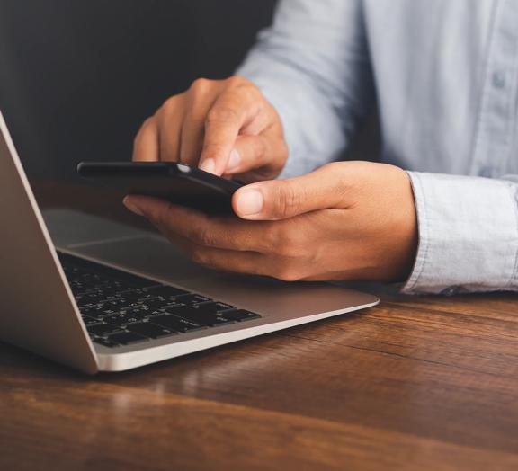 Close up of hands using a smartphone while working on a laptop placed on a wooden desk in a modern office setting.