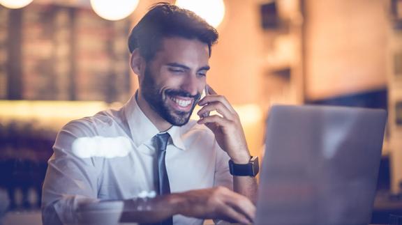 Professional man in a white shirt and tie smiling while talking on phone and working on a laptop in a modern indoor setting with warm lighting.