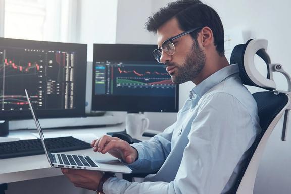 Person at desk analyzing financial charts on multiple monitors and a laptop, indicating a professional trading or financial analysis setup.