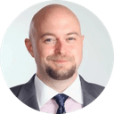 Portrait of a bald man with a short beard wearing a suit, dress shirt, and tie against a plain white background.