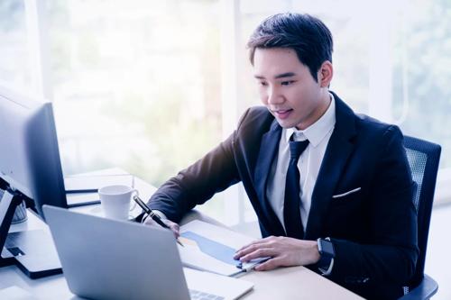 Business professional in a suit working at a desk with a laptop, desktop monitor, notepad, and coffee cup in a modern office setting