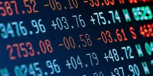 A digital screen showing colorful stock index numbers, price changes, and percentage variations in green, red, and orange on a dark background. The display represents market performance and trading data.