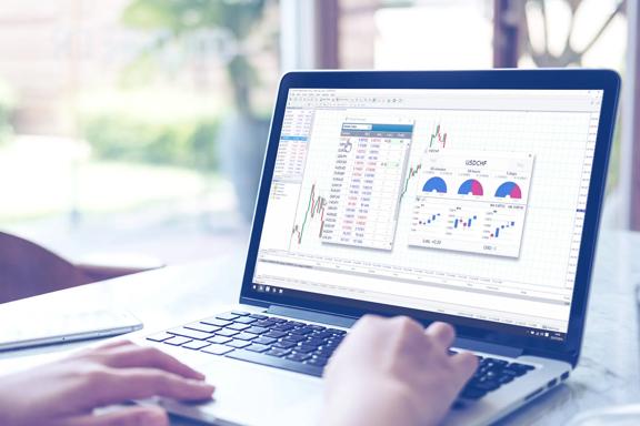 Person using a laptop displaying financial trading software with candlestick charts, currency pairs, and data indicators
