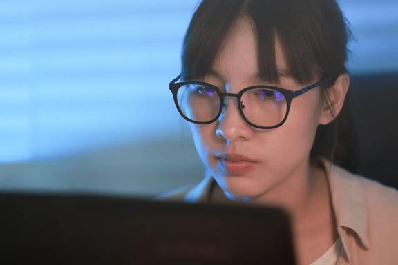 Close-up view of a person working on a laptop in a softly lit environment with horizontal blinds in the background