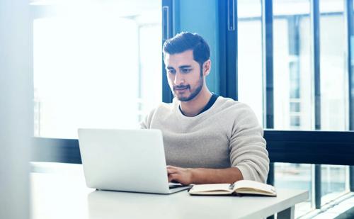 Man working on a laptop at a desk with an open notebook in a naturally lit office or study area