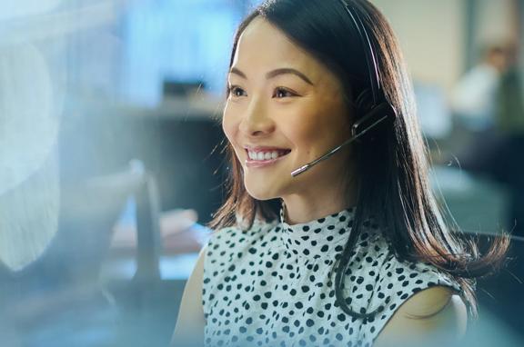 Woman wearing a headset and white shirt working at a desk in a modern office environment. Background shows other workstations and blurred figures, indicating a professional call center setting.